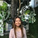 Photo of Chelina. She is wearing a dress and stands in front of a tree and hanging plants. 